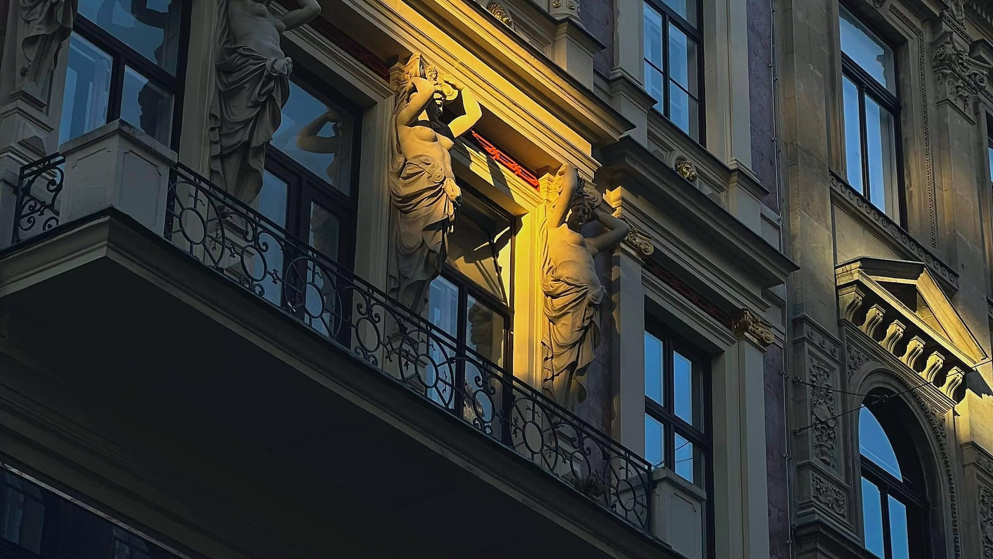 Fassade eines Wiener Zinshauses am Abend, mit Karyatiden, Balkon und einem erleuchteten Fenster.