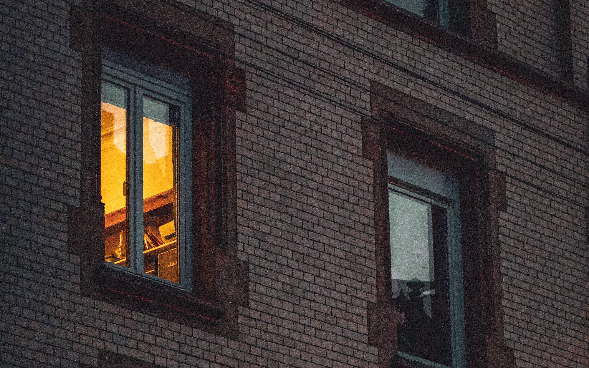 Altbaufassade aus Ziegel in der Dämmerung, vier Fenster, in einem davon brennt warmes Licht.