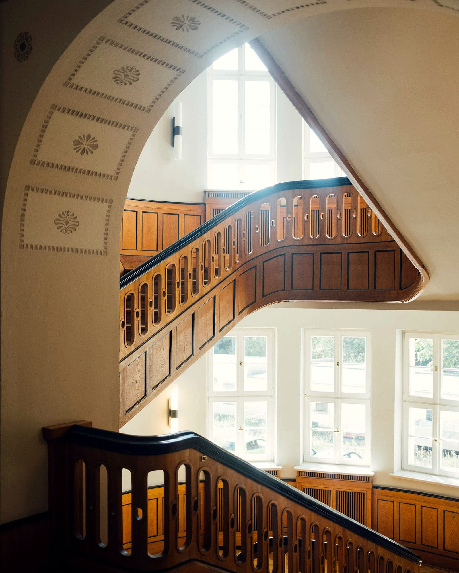 Blick in das Stiegenhaus eines Altbaus, Holzgeländer, stuckierter Bogen und hohe Fenster.